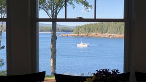 Vista desde una ventana sobre un lago con un bote, rodeado de árboles y una silla con un arreglo floral.