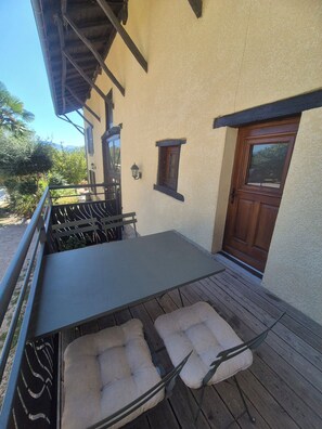 Ein überdachter Außenbereich mit Tisch und gepolsterten Stühlen, einer Holzterrasse und einem Gebäude mit Tür und Fenstern.
