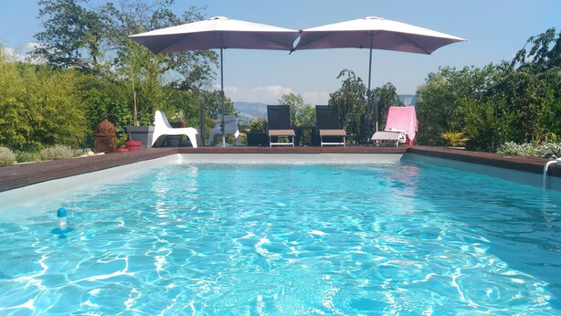 A swimming pool with clear blue water, surrounded by lounge chairs and umbrellas, with greenery and a clear sky in the background.