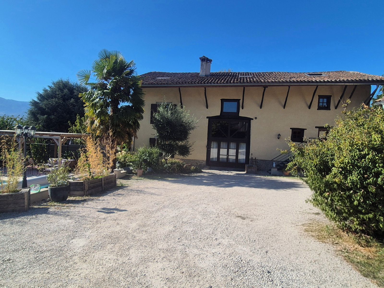 A building with a gravel driveway leading to a gated entrance, surrounded by greenery and a clear sky.