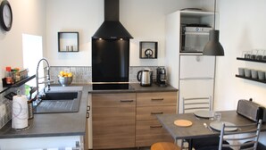 A modern kitchen with a black range hood, a built-in oven, and a coffee maker. There is a dining table with chairs and a small island with a sink.