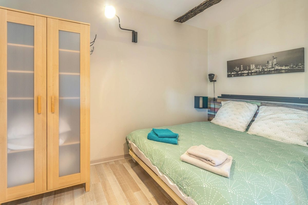 A bedroom with a bed, a wardrobe, a cityscape painting, and a wall-mounted light.