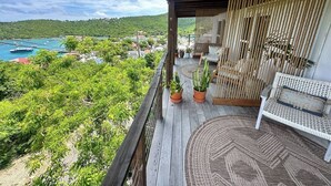 Un balcón de madera con un sofá de mimbre y dos plantas en macetas, con vistas a un paisaje verde exuberante y una masa de agua.