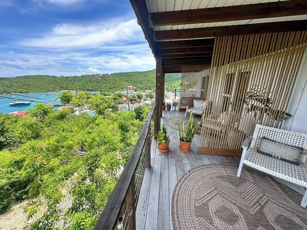 A wooden balcony with a wicker sofa and two potted plants, overlooking a lush green landscape and a body of water.
