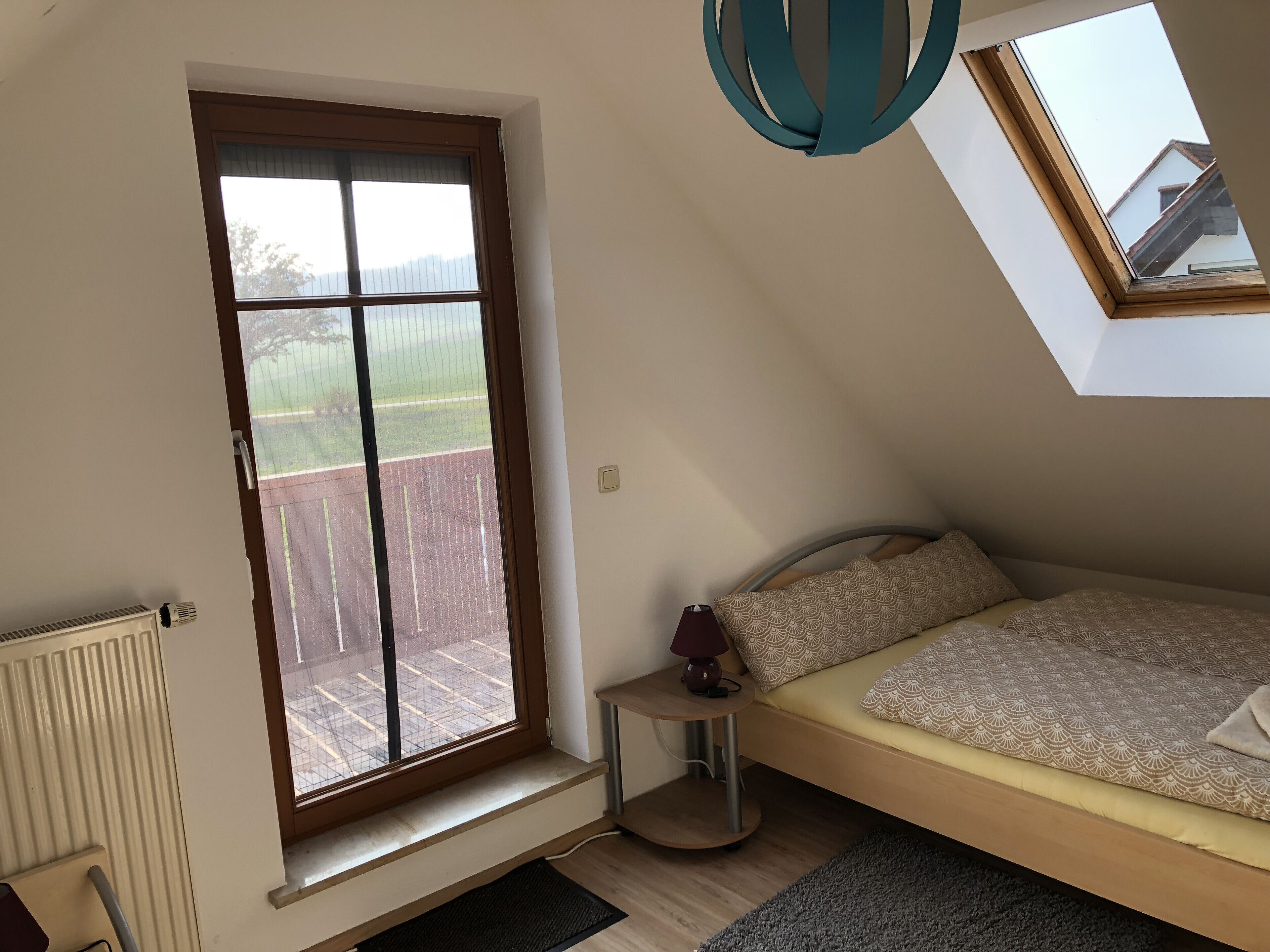 Ein Schlafzimmer mit Oberlicht, einem Bett mit gemusterten Betttextilien, einem Nachttisch mit Lampe und Blick auf eine Holzterrasse durch ein Fenster.