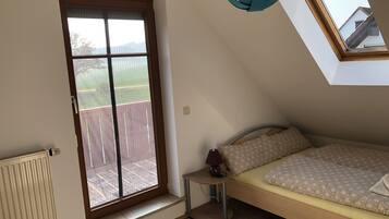 Ein Schlafzimmer mit Oberlicht, einem Bett mit gemusterten Betttextilien, einem Nachttisch mit Lampe und Blick auf eine Holzterrasse durch ein Fenster.