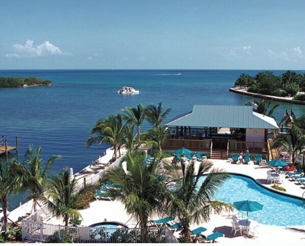 Relax by the pool surrounded by lounge chairs and umbrellas, with a boat on the water and palm trees nearby.