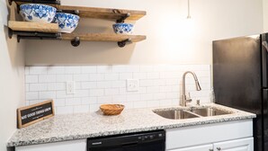 A kitchen with white subway tiles, a granite countertop, a stainless steel sink, a black dishwasher, and wooden shelves holding blue and white bowls. - Downtown Greenville, SC Southern Simplicity! (Greenville)