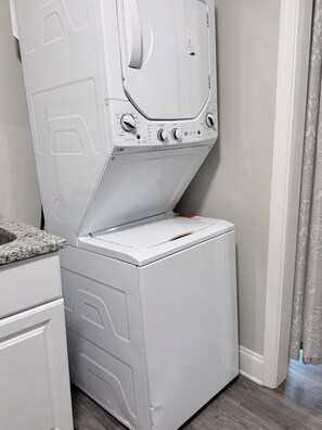 A compact, white front-loading washing machine with a control panel, situated in a small laundry room with a wooden floor and a patterned curtain.