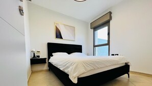 Un dormitorio moderno con estructura de cama negra, ropa de cama blanca, una mesita de noche con lámpara y un ventanal que proporciona luz natural.