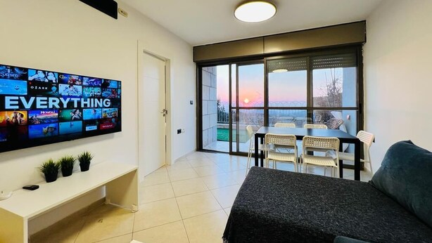 Un salón moderno con un televisor de pantalla plana mostrando 'EVERYTHING' en la pantalla, un sofá, una mesa de comedor con sillas y un balcón con vistas al atardecer.