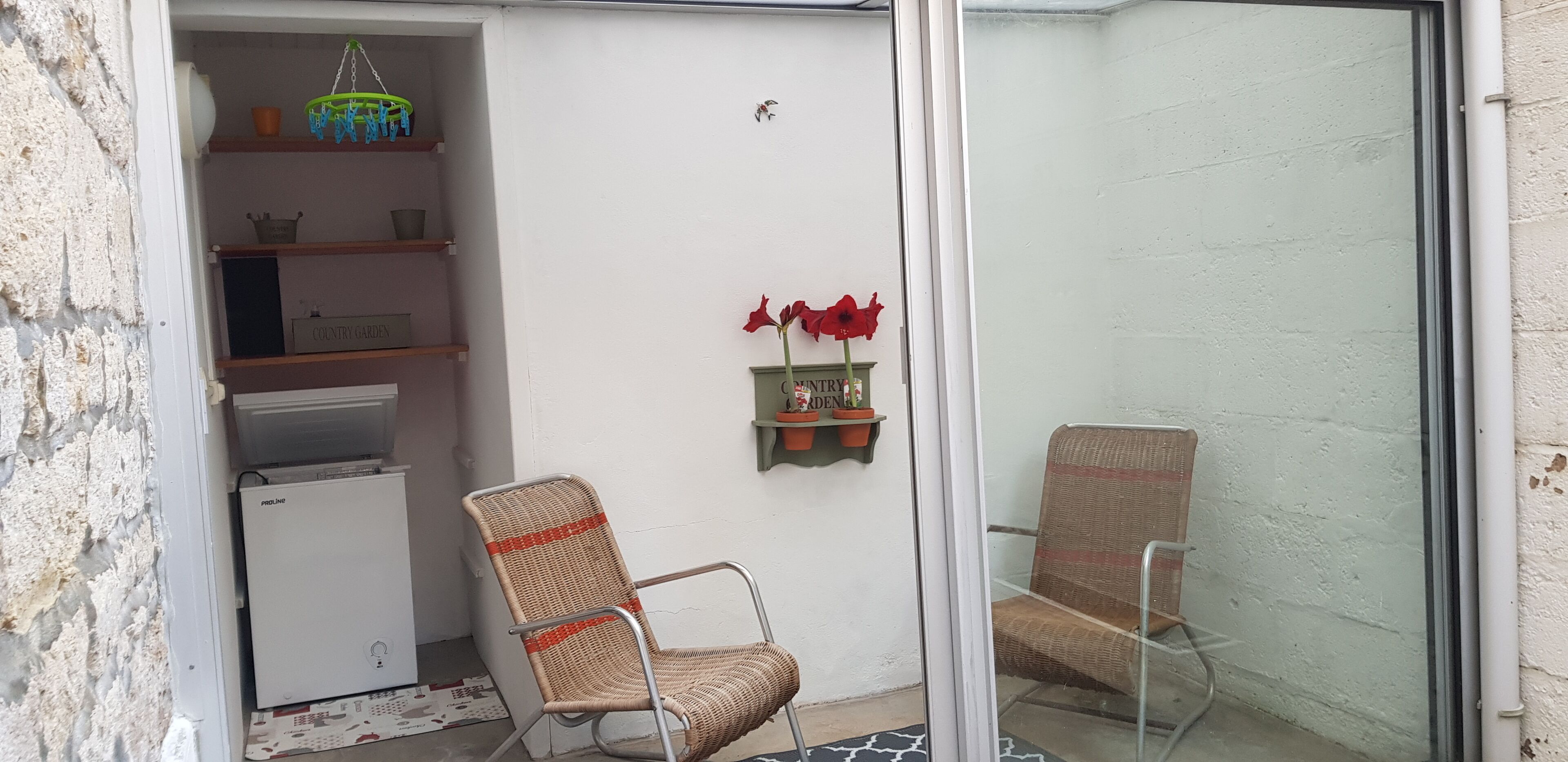 Quarto pequeno com parede branca, uma cadeira de vime, radiador branco e uma pequena prateleira com flores vermelhas.