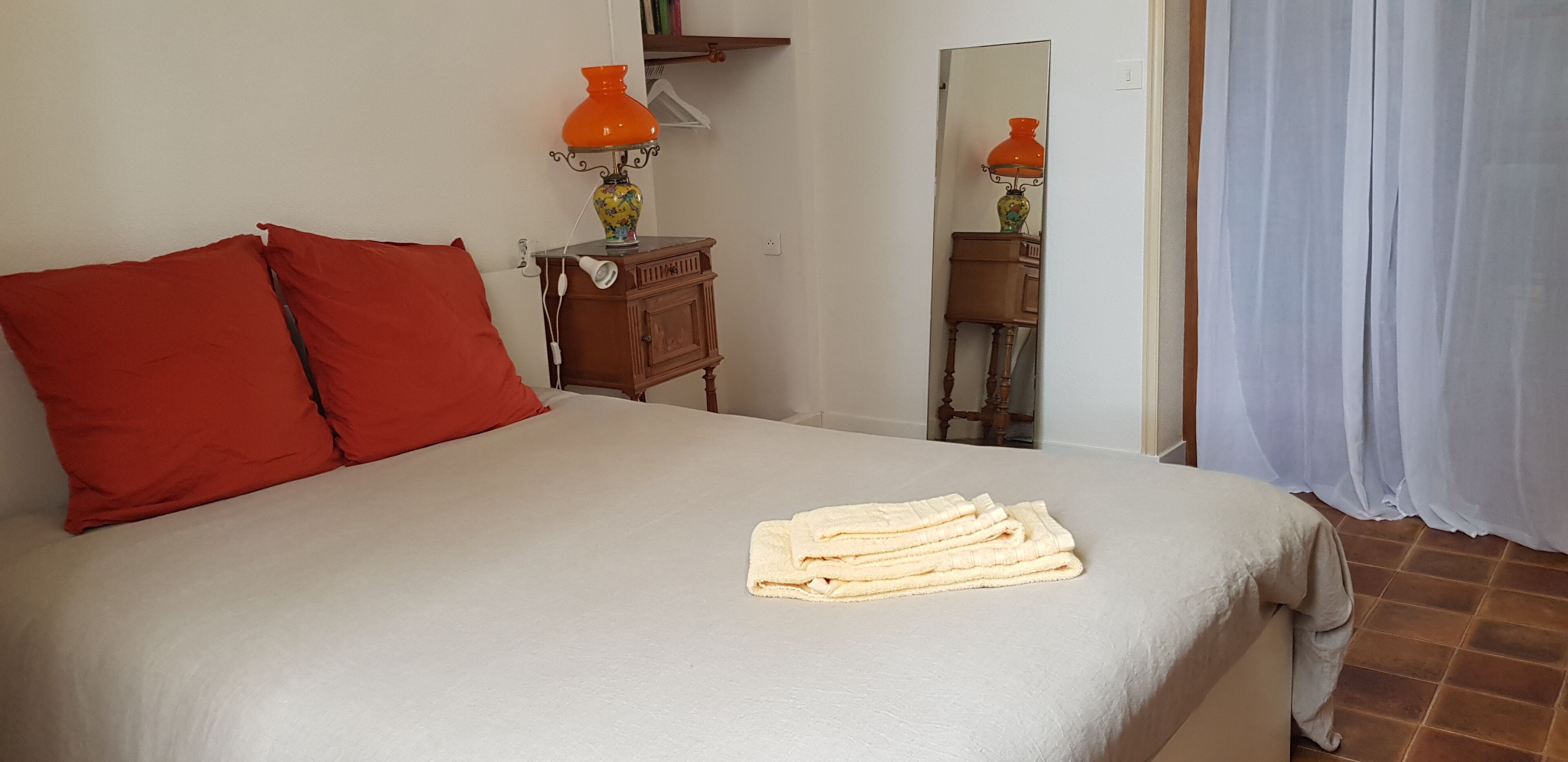 A bed with red pillows, a white blanket, and a folded towel. There is a small wooden nightstand with an orange lamp and a vase. A mirror reflects part of the room.
