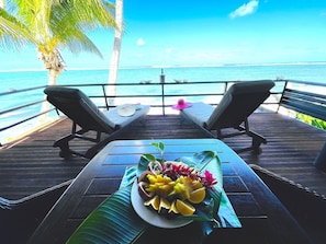 A wooden deck with two lounge chairs facing a beach, a table with fruit and flowers, and a palm tree. - Vaka Rua - right over the golden sand facing the lagoon with air con Free WIFI (Vaimaanga)