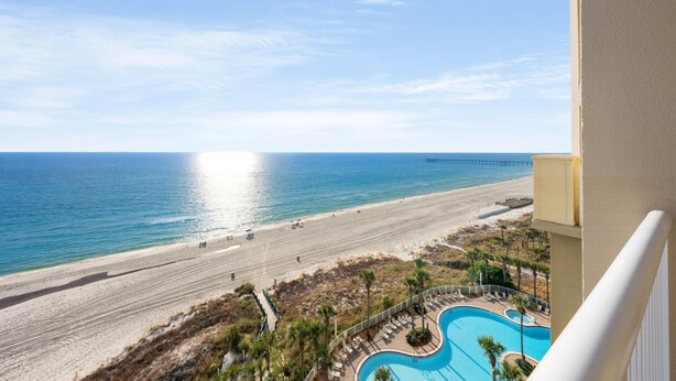 A beach view from a balcony with a swimming pool and a pier in the distance.