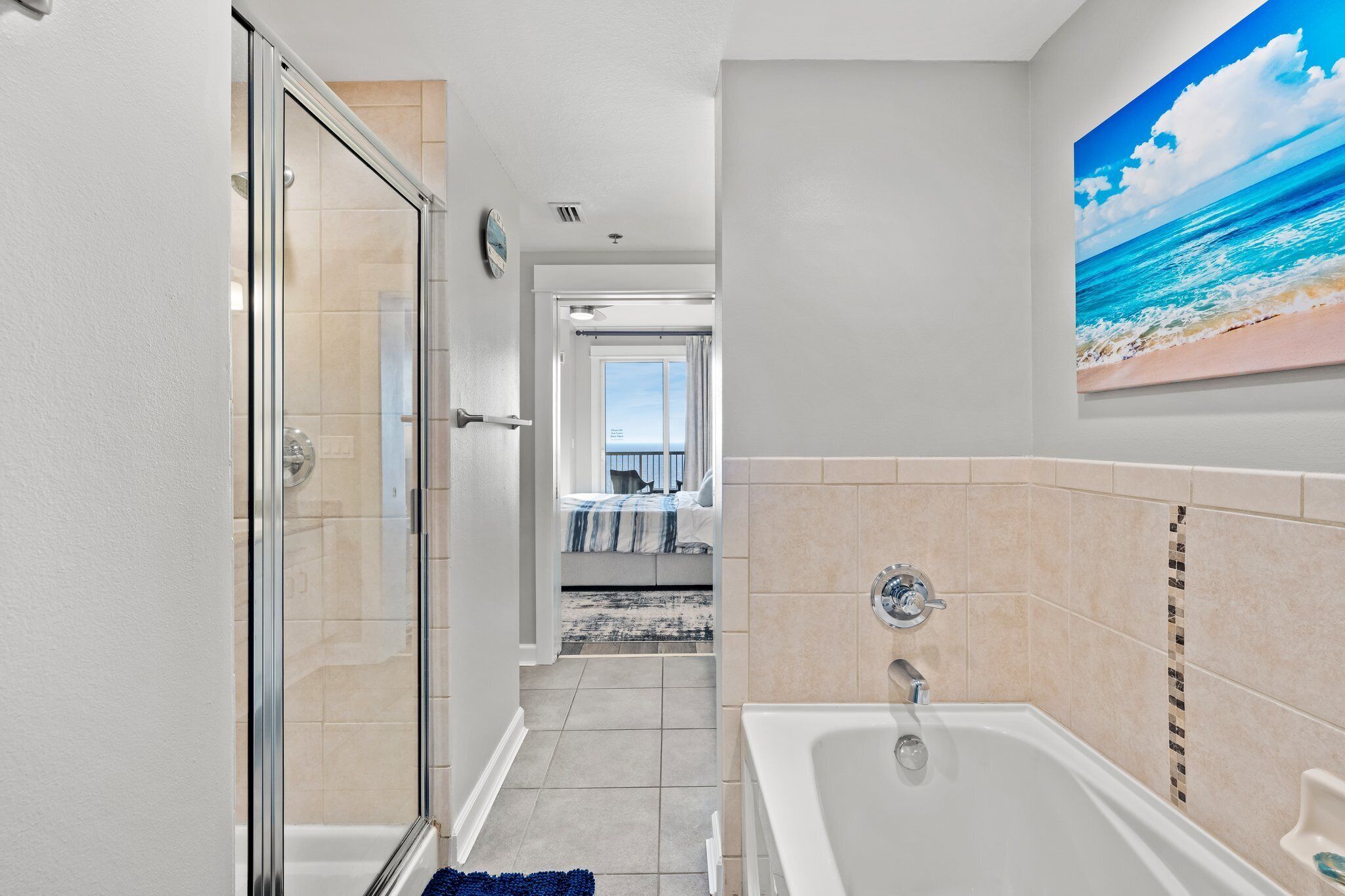 A bathroom with a glass shower door, a bathtub, and a beach-themed painting on the wall.