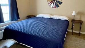 A bedroom with a large bed, a wall clock, a window with curtains, and a nightstand with a lamp. - Hunter's Ridge Cabins - 2 (Concan)