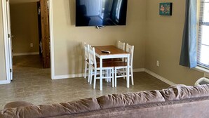 A living room with a TV mounted on the wall, a dining table with chairs, and a brown couch. - Hunter's Ridge Cabins - 2 (Concan)