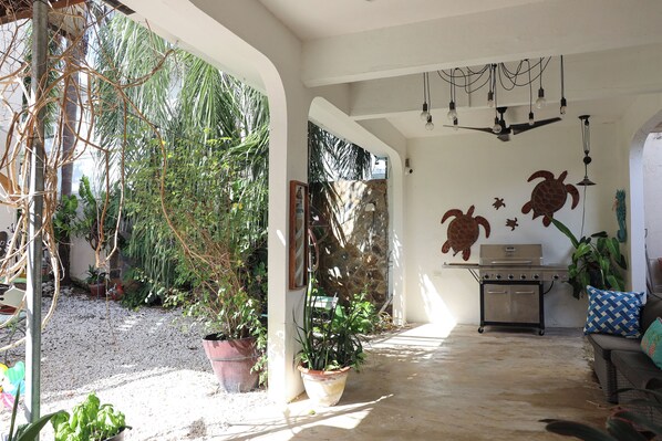 A covered patio area with a grill, potted plants, and a seating area with a patterned cushion.