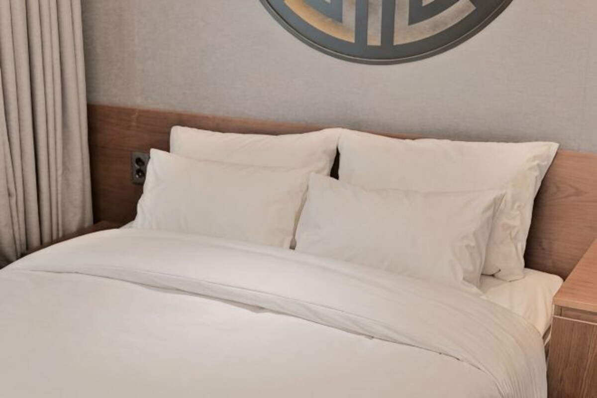 A bed with white bedding, a wooden headboard, and a wall-mounted light fixture.
