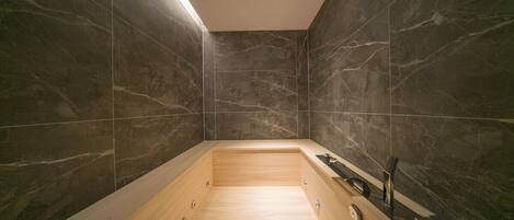 A modern bathroom with dark marble walls, a wooden bathtub, and a sleek black faucet.