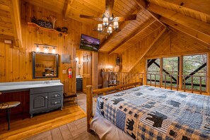 Ein Schlafzimmer in einer Blockhütte mit einem großen Bett, Holzwänden, einem Deckenventilator, einem angebrachten Fernseher und Blick ins Freie.