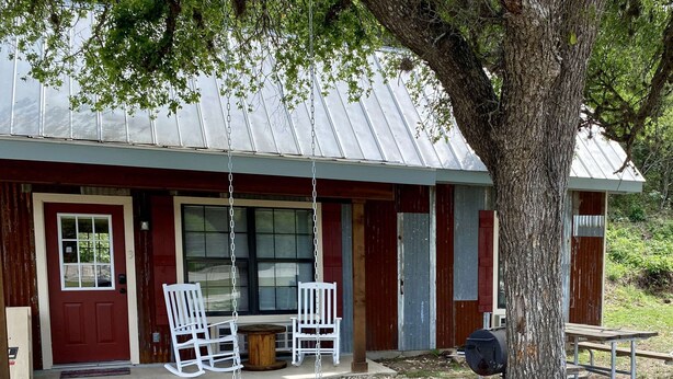 A rustic cabin with a porch, two white chairs, a swing, and a wooden table. - Hunter's Ridge Cabins - 3 (Concan)