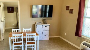 A living room with a brown leather couch, a wooden dining table with white chairs, a white cabinet, a flat-screen TV mounted on the wall, and a window with a red curtain. - Hunter's Ridge Cabins - 3 (Concan)
