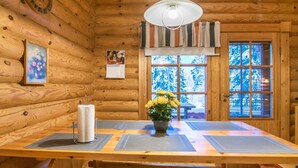 Interior de una cabaña de madera con una mesa, una lámpara colgante, una ventana que muestra árboles nevados y un cuadro en la pared.