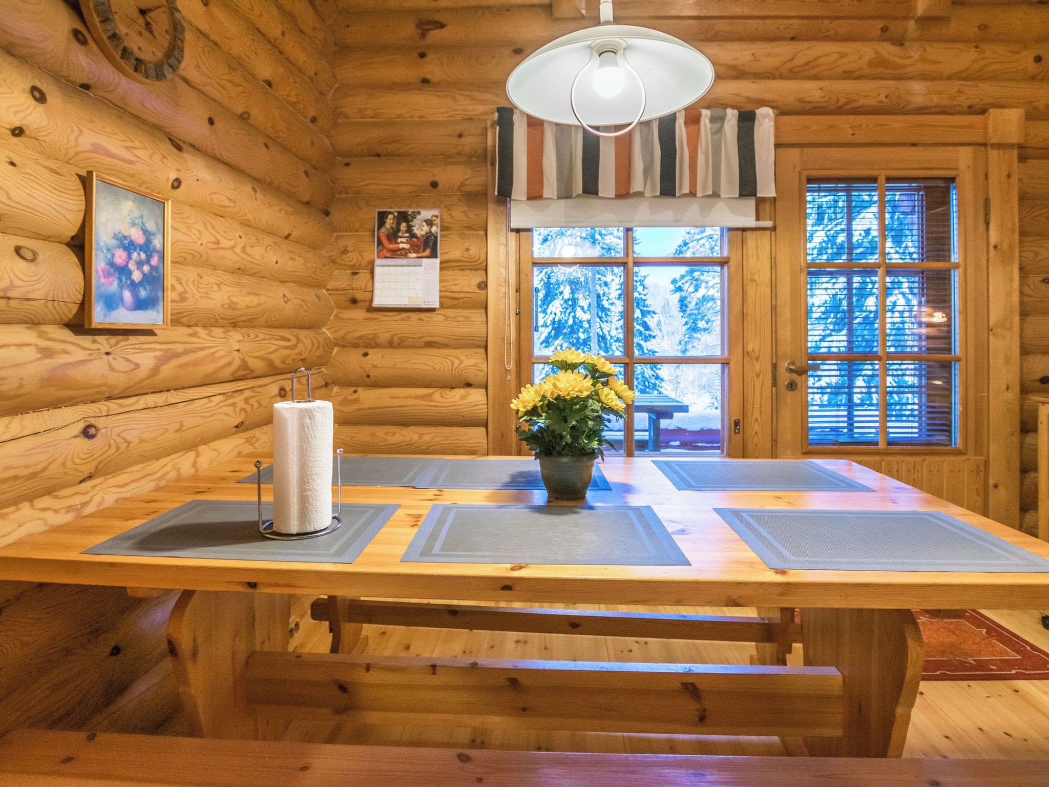 Interior de una cabaña de madera con una mesa de comedor, bancos, una lámpara colgante y una ventana que ofrece vistas de árboles cubiertos de nieve.