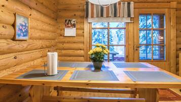 Interior de una cabaña de madera con una mesa de comedor, bancos, una lámpara colgante y una ventana que ofrece vistas de árboles cubiertos de nieve.