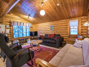 Interior de una cabaña de troncos con sofá, sillas, televisor y chimenea.