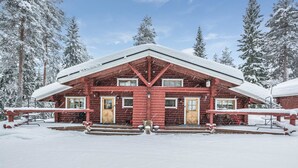 Una cabaña de troncos rojos con techo nevado, puertas numeradas y un porche con bancos.