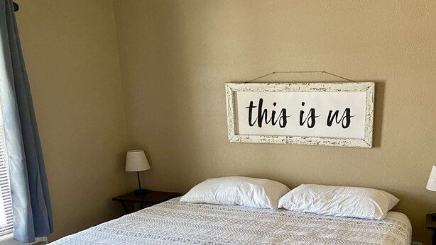 A neatly made bed with a patterned bedspread, a wall-mounted sign saying "this is us", and a window with curtains.