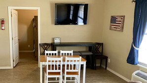 A living room with a brown couch, a dining table with white chairs, a TV mounted on the wall, and a flag decoration.