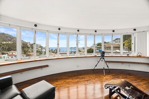Quarto espaçoso com grandes janelas que oferecem vista para o mar e edifícios ao redor.
