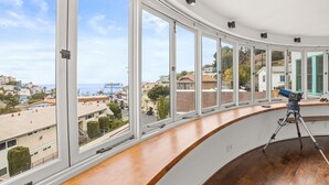 Una habitación con grandes ventanales que dan a vistas de un pueblo costero, un telescopio sobre una plataforma de madera y un techo curvo.