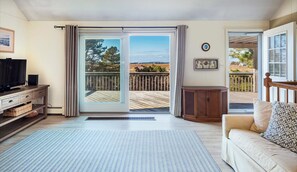 A living room with a TV, a sofa, a wooden cabinet, and a view of a deck and landscape through a sliding glass door. - Sunset Views - Cape Cod Bay. (Eastham)