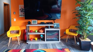 A living room with a large flat-screen TV mounted on an orange wall, two yellow chairs, a small plant, and a colorful rug.