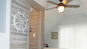 Une chambre avec une porte en bois, un ventilateur de plafond, un lit et une fenêtre avec des stores.