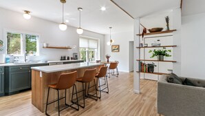 Eine moderne Küche mit Kochinsel, Barhocker und Pendelleuchten. Es gibt einen Essbereich mit Tisch und Stühlen sowie eine Wohnbereitschaft mit Sofa und Bücherregal.