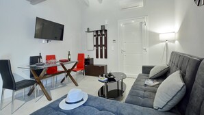 A modern living room with a grey sofa, a glass dining table with red chairs, a flat-screen TV mounted on the wall, and a white door.