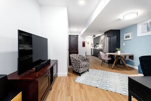 Un soggiorno moderno con una TV a schermo piatto, una poltrona decorata, un tavolo da pranzo e una cucina sullo sfondo.