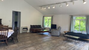 A spacious living area with a high ceiling, wooden flooring, a sofa, a chair, a dining table with chairs, a television, and large windows offering a view of greenery.