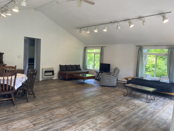 A spacious living area with a high ceiling, wooden flooring, a sofa, a chair, a dining table with chairs, a television, and large windows offering a view of greenery.