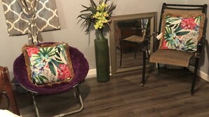 Ein Wohnzimmer mit einem violetten Sessel, einem Holzstuhl mit einem blumigen Kissen, einem violetten Teppich, einer grünen Vase mit Blumen und einem Spiegel an der Wand.