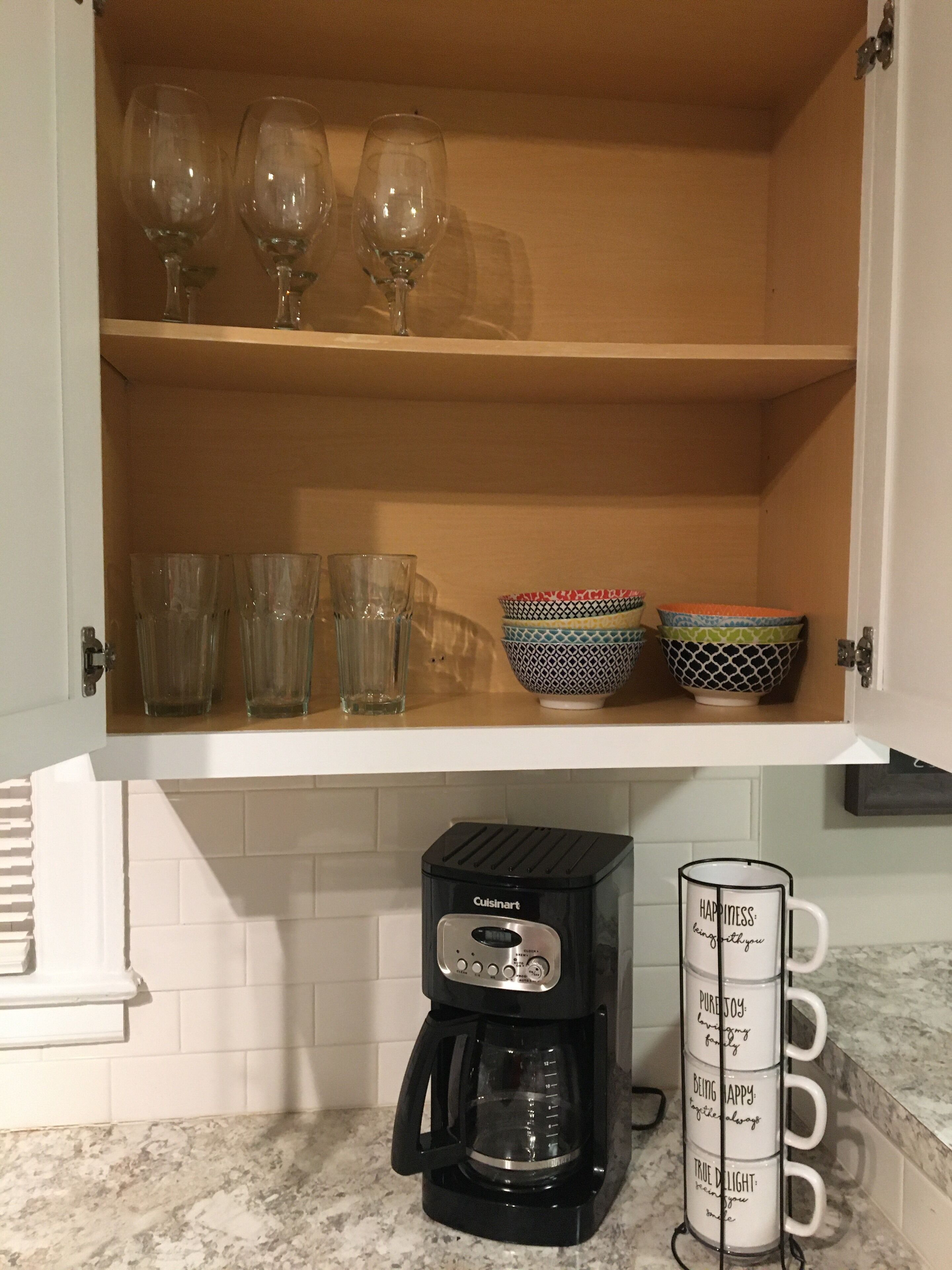 A kitchen with a built-in shelf holding wine glasses and bowls, a Cuisinart coffee maker, and a mug rack with motivational phrases.