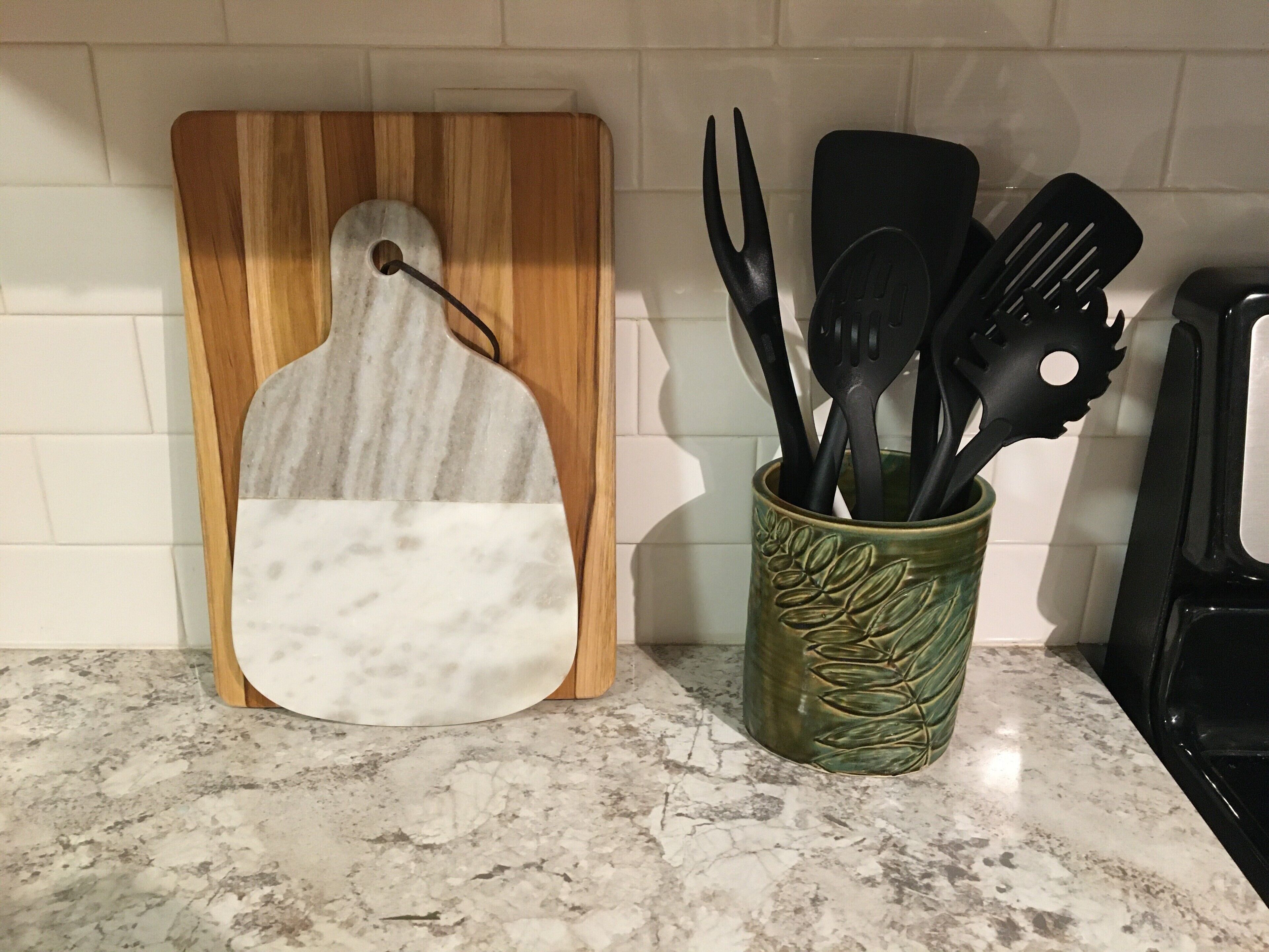 A kitchen counter with a wooden cutting board, a marble backsplash, and a green utensil holder.