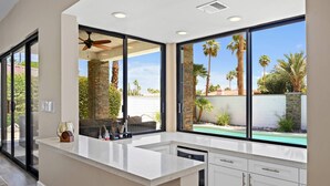 A modern kitchen with large windows, a built-in dishwasher, and a view of a swimming pool and palm trees.