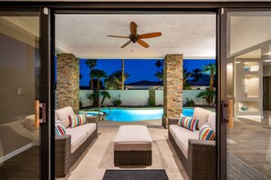 Ein moderner Außenbereich mit Pool, zwei Korbsessel-Sofas mit bunten Kissen, einem Deckenventilator und Blick auf Palmen und ein Gebäude im Hintergrund.